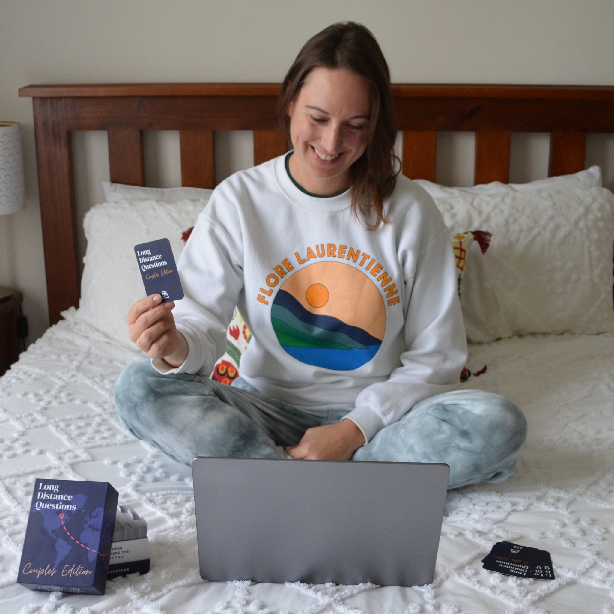 Person sitting on bed, smiling, holding a card labeled 'Long Distance Questions' next to a laptop.